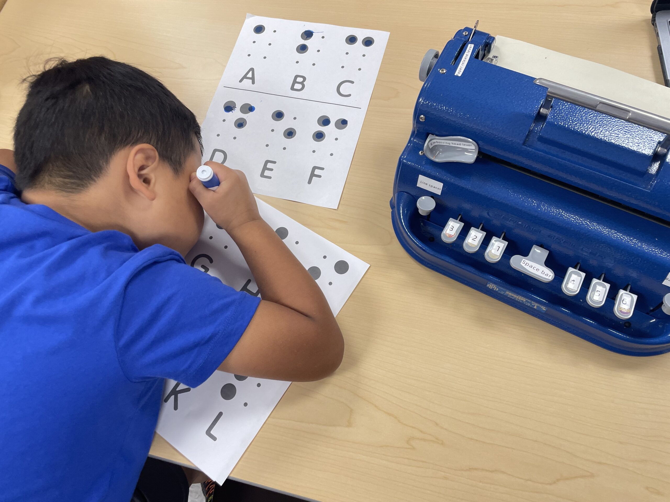 Young male student using a dot marker to fill in a large braille alphabet worksheet. His blue Perklns Brailler is on the table to the right of him.